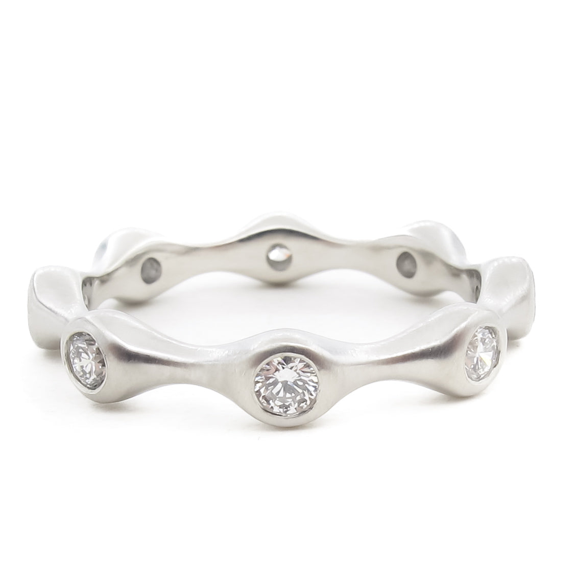 A platinum or white gold eternity ring with an undulating wave design. The band features bezel-set round brilliant diamonds positioned at the peaks of the wave pattern. The metal has a satin finish that contrasts with the sparkle of the diamonds. The ring is photographed straight-on against a white background.