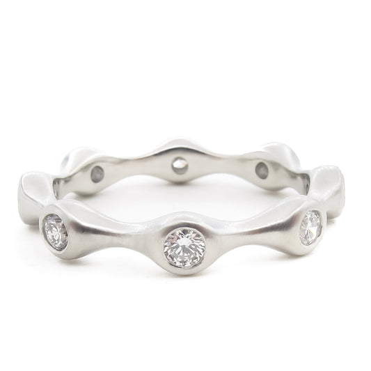 A platinum or white gold eternity ring with an undulating wave design. The band features bezel-set round brilliant diamonds positioned at the peaks of the wave pattern. The metal has a satin finish that contrasts with the sparkle of the diamonds. The ring is photographed straight-on against a white background.