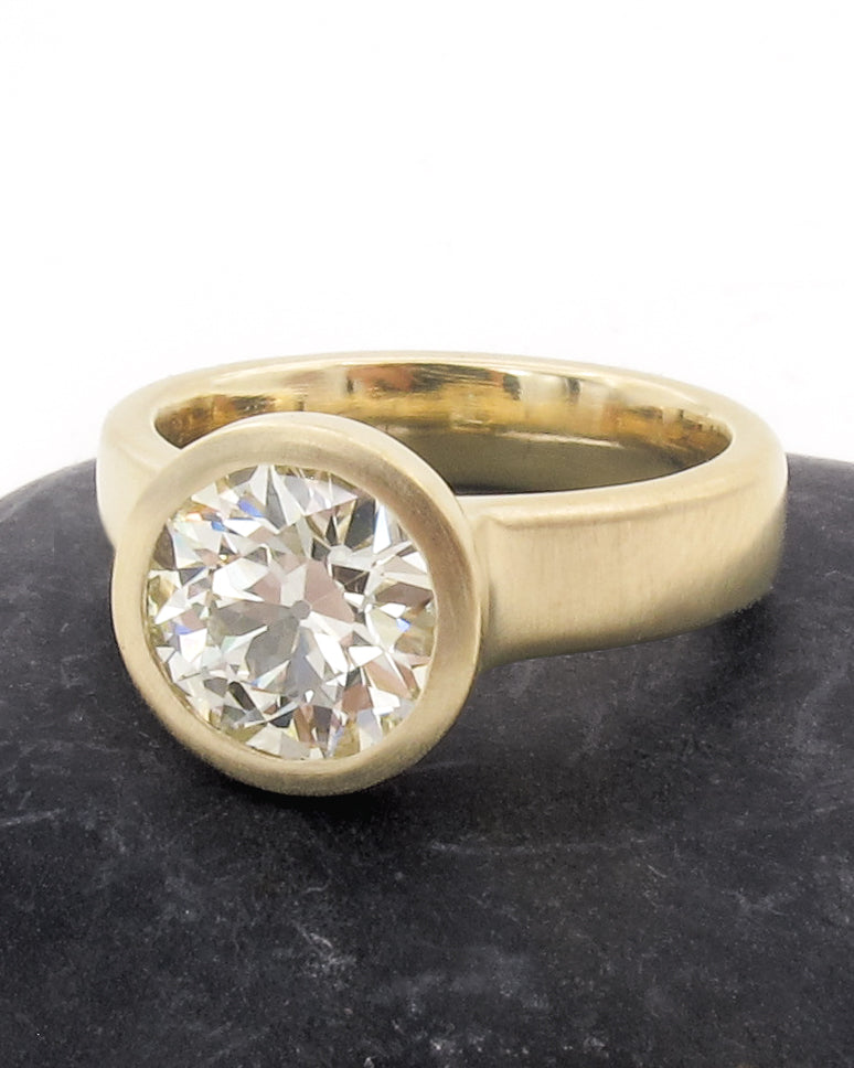 A yellow gold ring with a large round brilliant-cut diamond set in a bezel setting, resting on a dark textured surface against a white background.